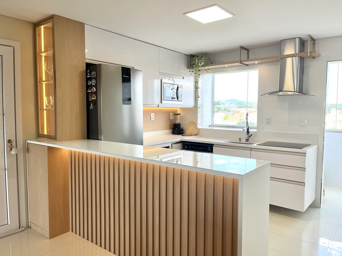 Cozinha planejada em branco e madeira clara, com bancada em U, painel ripado e cristaleira iluminada